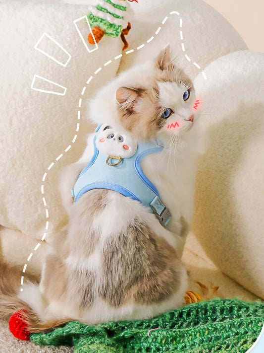 Cat wearing a blue harness with a cartoon design on a soft surface.