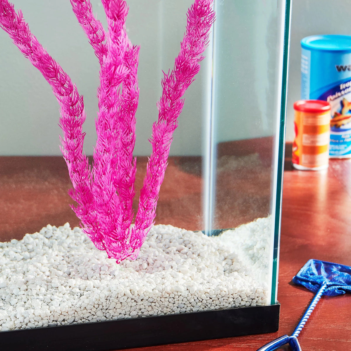 Small aquarium with pink artificial plants and white gravel on a wooden surface.